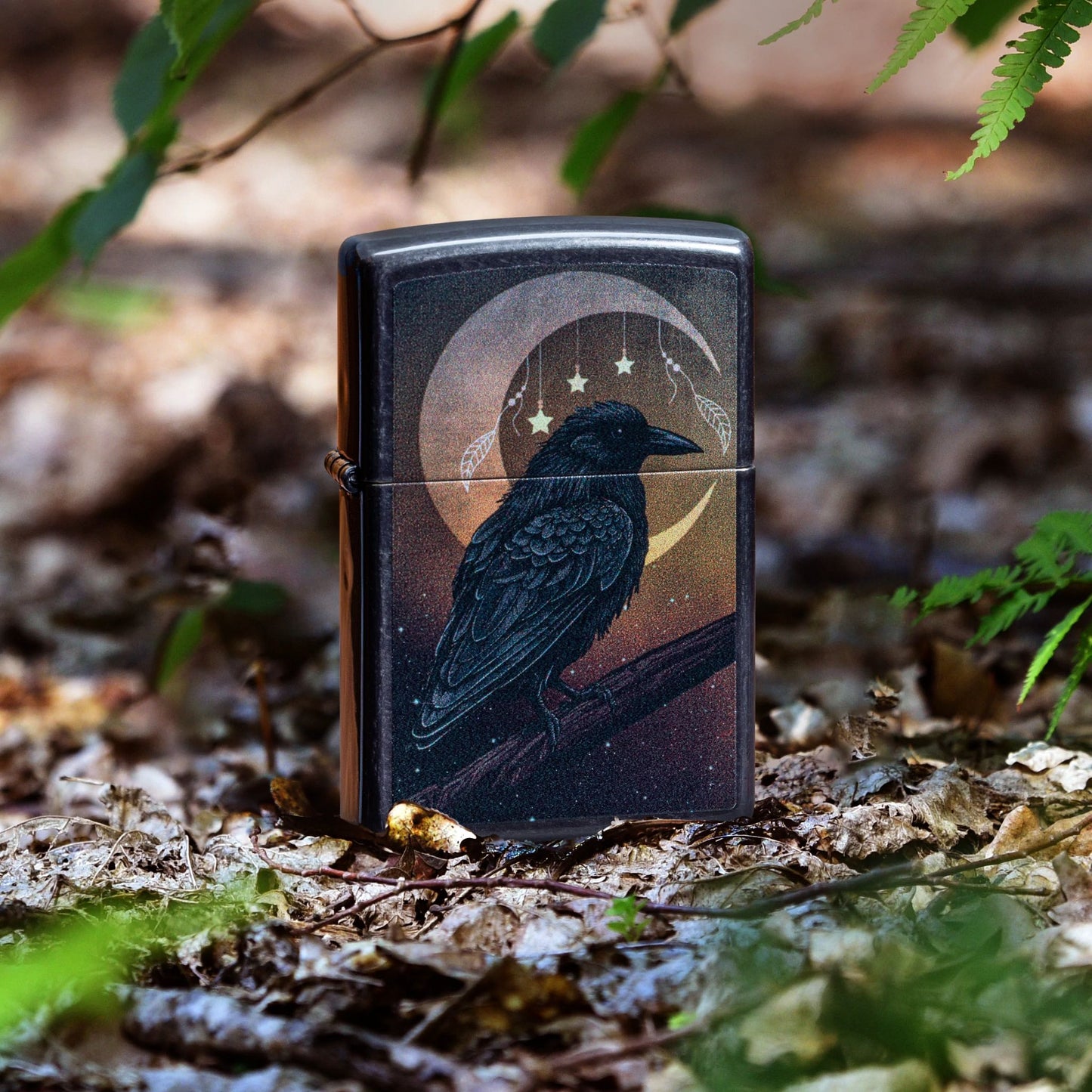 Lifestyle image of Zippo Raven Moon Design Grey Windproof Lighter standing on the forest floor showing the front of the lighter with green leaves around the lighter. 

