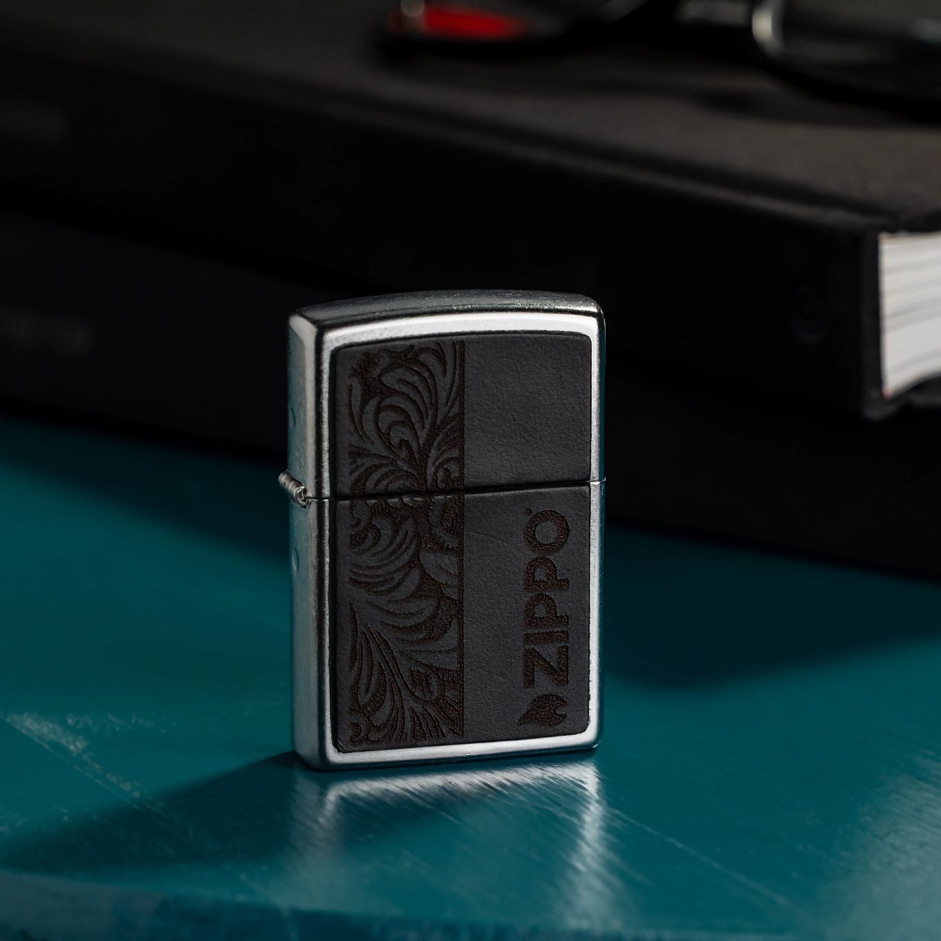 Lifestyle image of Zippo Black Leather Emblem - Zippo Design Street Chrome Windproof Lighter standing on a desk next to books and a pair of eyeglasses.

