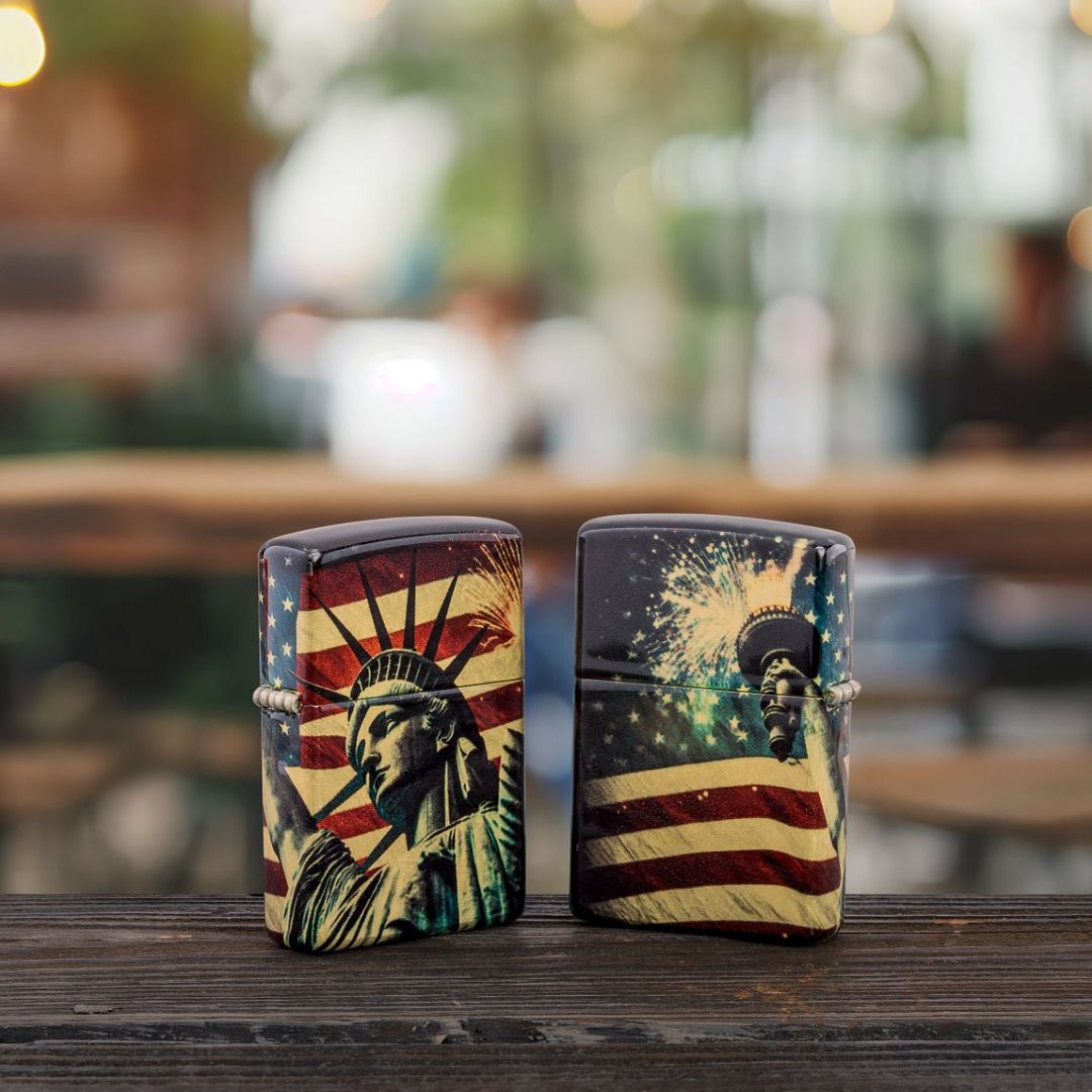 Lifestyle image of two Zippo Spark of Liberty Design 540 Tumbled Brass Windproof Lighters; one showing the front and hinge side of the lighter, while the other shows the back of the lighter. They are both standing on a wooden surface with an out of focus restaurant as the background. 


