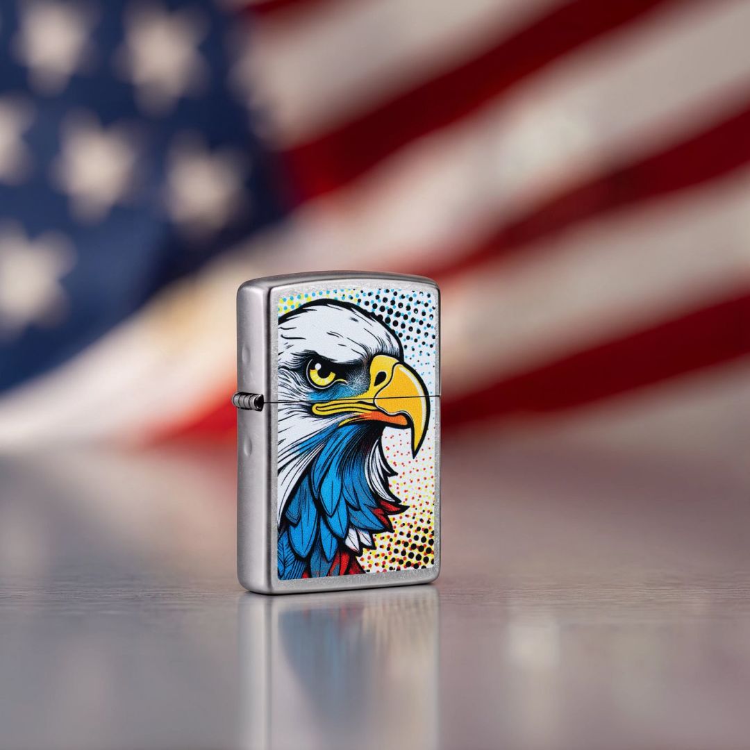 Lifestyle image of Zippo Pop Art Eagle Design Street Chrome Windproof Lighter standing on a grey surface showing the front and hinge side of the lighter with an out of focus American flag as the background. 


