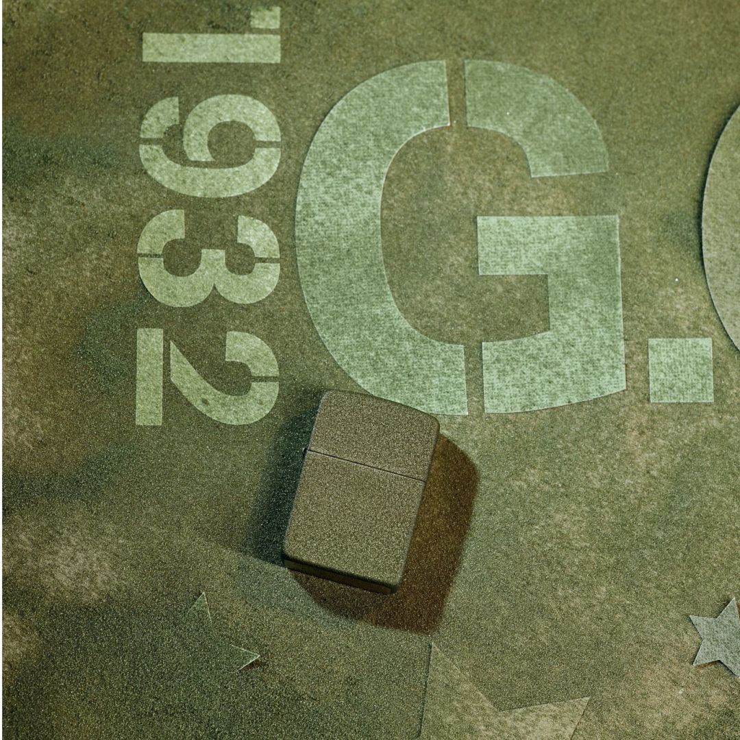 Lifestyle image of Zippo 2026 Founder’s Day Commemorative Windproof Lighter lying on a camouflage green surface showing the front of the lighter. The background is camouflage green with 1932, a G and stars in a lighter green. 
