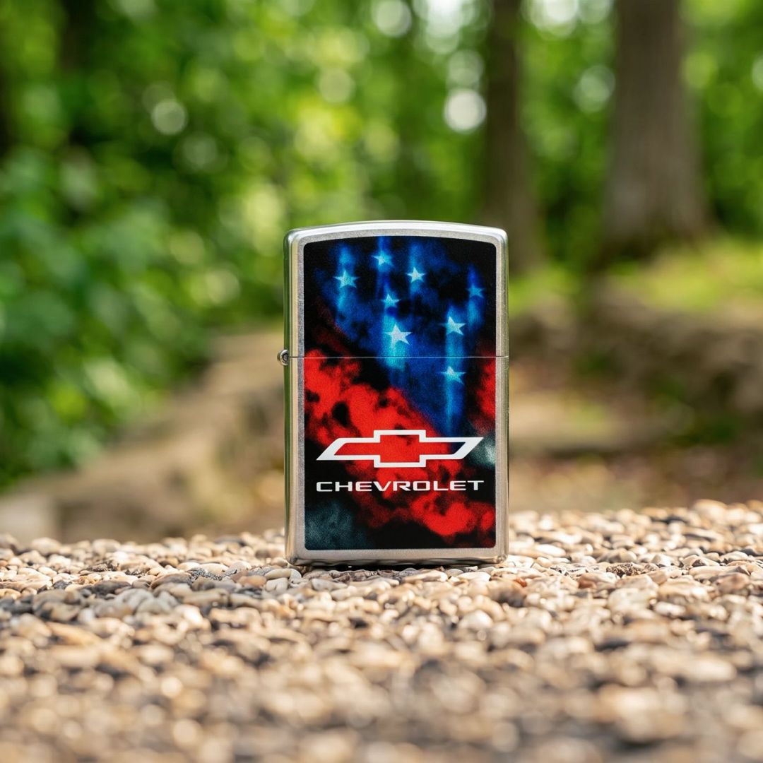Lifestyle image of Zippo Chevrolet® Flag Design Street Chrome Windproof Lighter standing on a gravel trail showing the front of the lighter with an out of focus nature background. 

