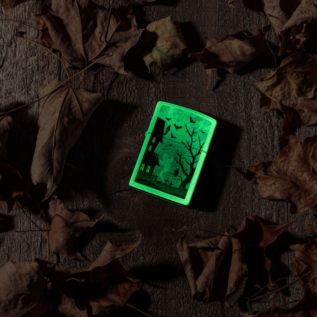 Lifestyle image of Zippo Spooky Design Glow in the Dark Green Windproof Lighter laying on a wooden surface surrounded by brown leaves while glowing in the dark.

