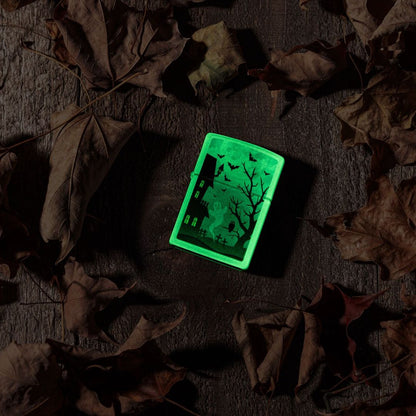 Lifestyle image of Zippo Spooky Design Glow in the Dark Green Windproof Lighter laying on a wooden surface surrounded by brown leaves while glowing in the dark.

