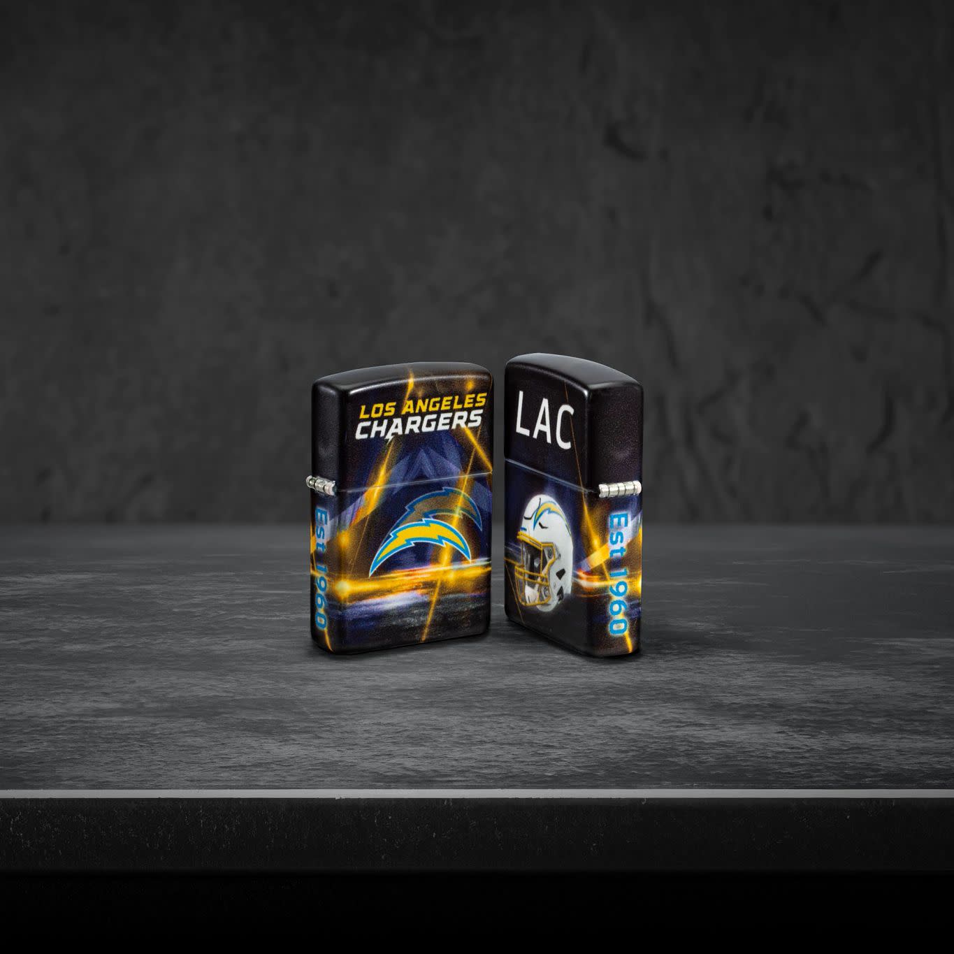 Lifestyle image of two Zippo NFL Los Angeles Chargers 540 Matte Windproof Lighters, one showing the front of the lighter and the other showing the back, standing on a dark grey surface.