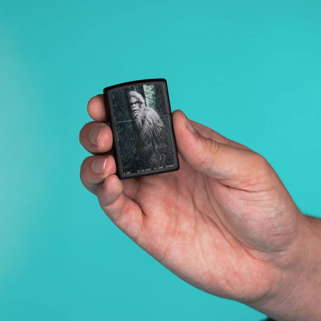 Lifestyle image of Zippo Squatch Cam Design Black Matte Windproof Lighter lit in hand showing the front of the lighter with turquoise-colored background.

