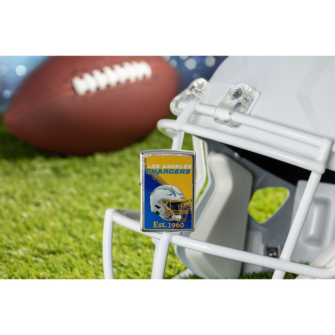 Lifestyle image of Zippo NFL Los Angeles Chargers Helmet Design Street Chrome Windproof Lighter standing on the facemask of a white helmet showing the front of the lighter with a football lying on a football field as the background.

