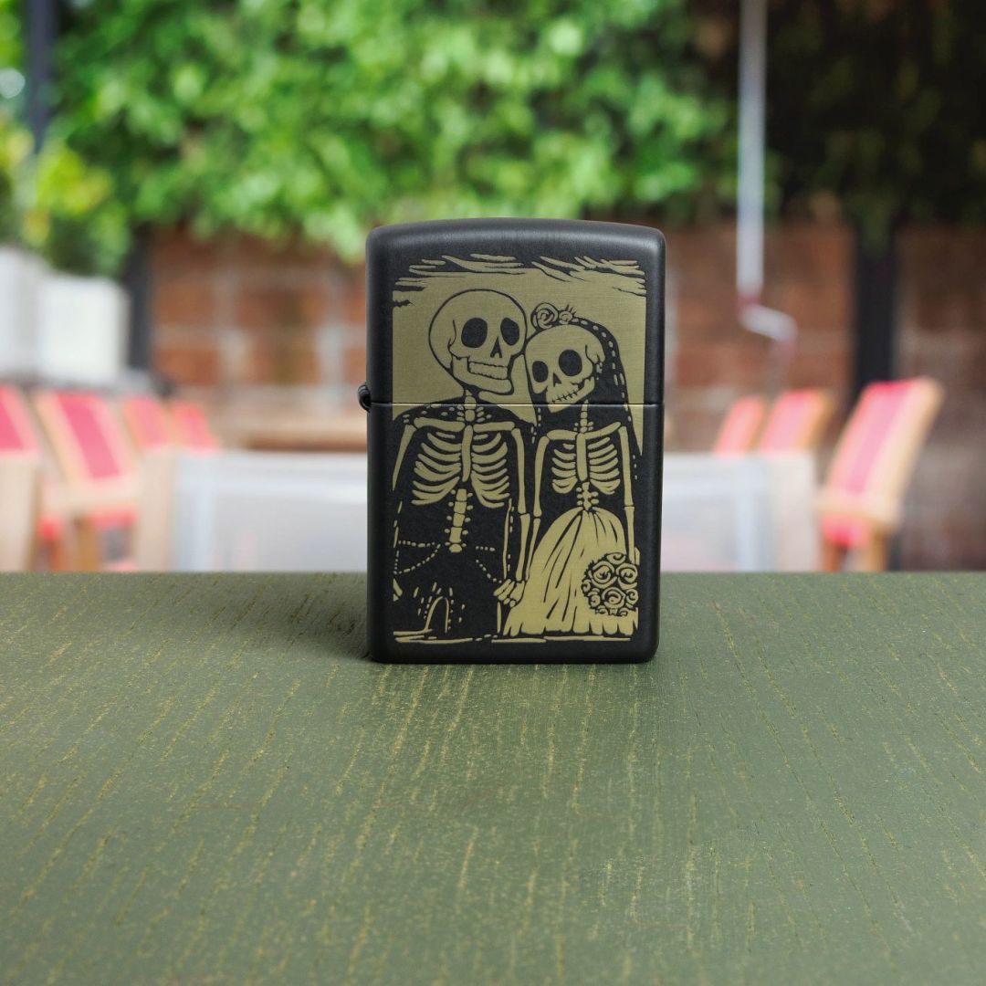 Lifestyle image of Zippo Skeleton Wedding Design Black Matte Windproof Lighter standing on a green wooden surface with an out of focus outdoor restaurant as the background with a brick wall and trees. 

