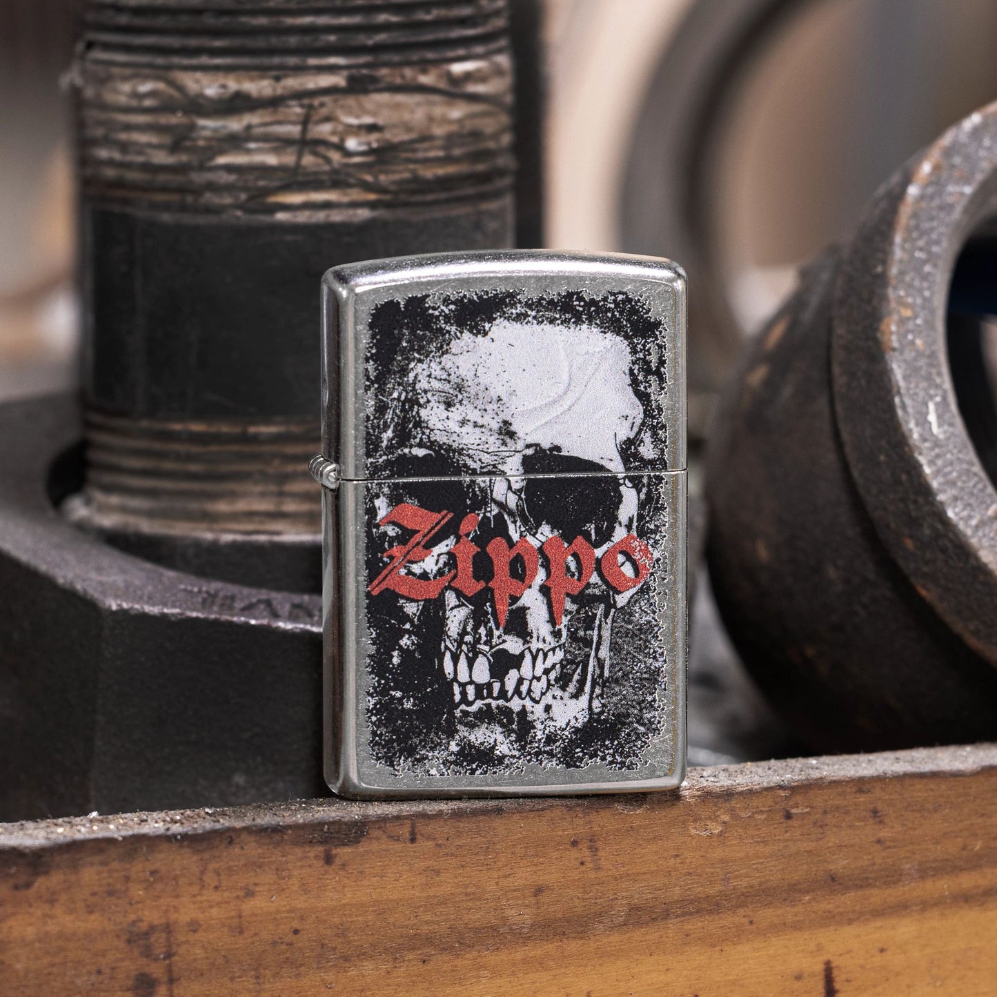 Lifestyle image of Zippo Skull Design Street Chrome Windproof Lighter standing on a workbench in front of pipe fittings while showing the front of the lighter. 


