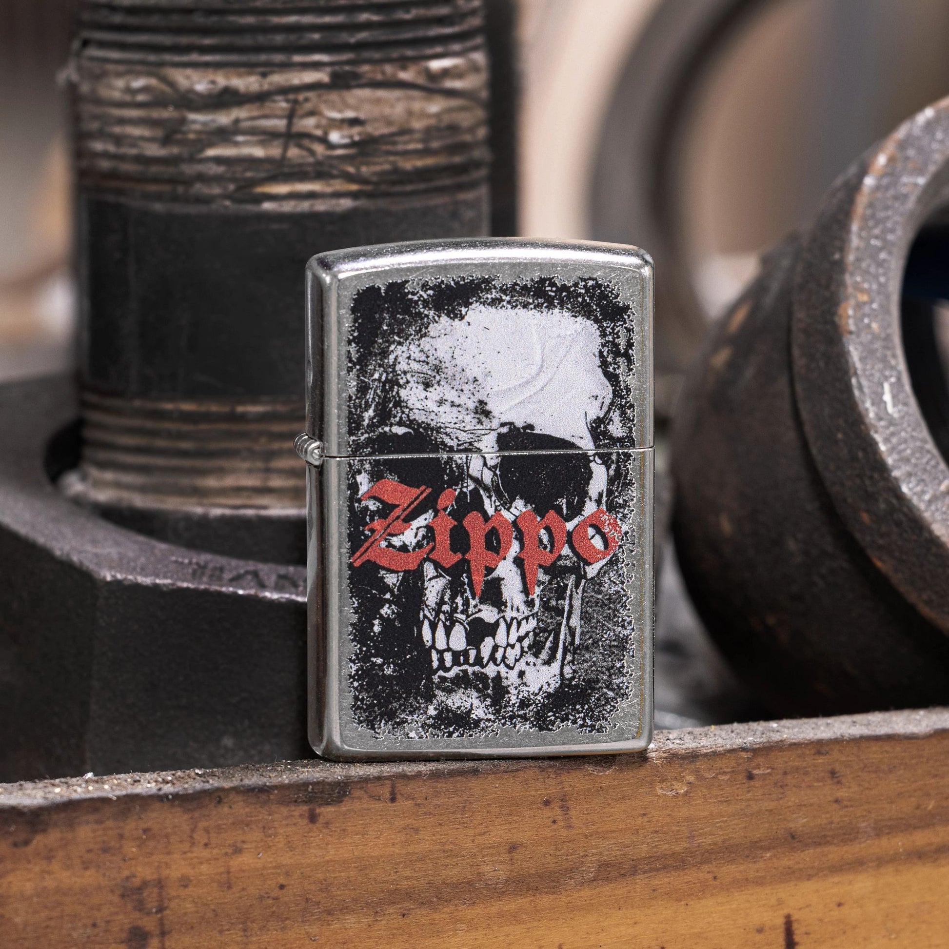 Lifestyle image of Zippo Skull Design Street Chrome Windproof Lighter standing on a workbench in front of pipe fittings while showing the front of the lighter. 


