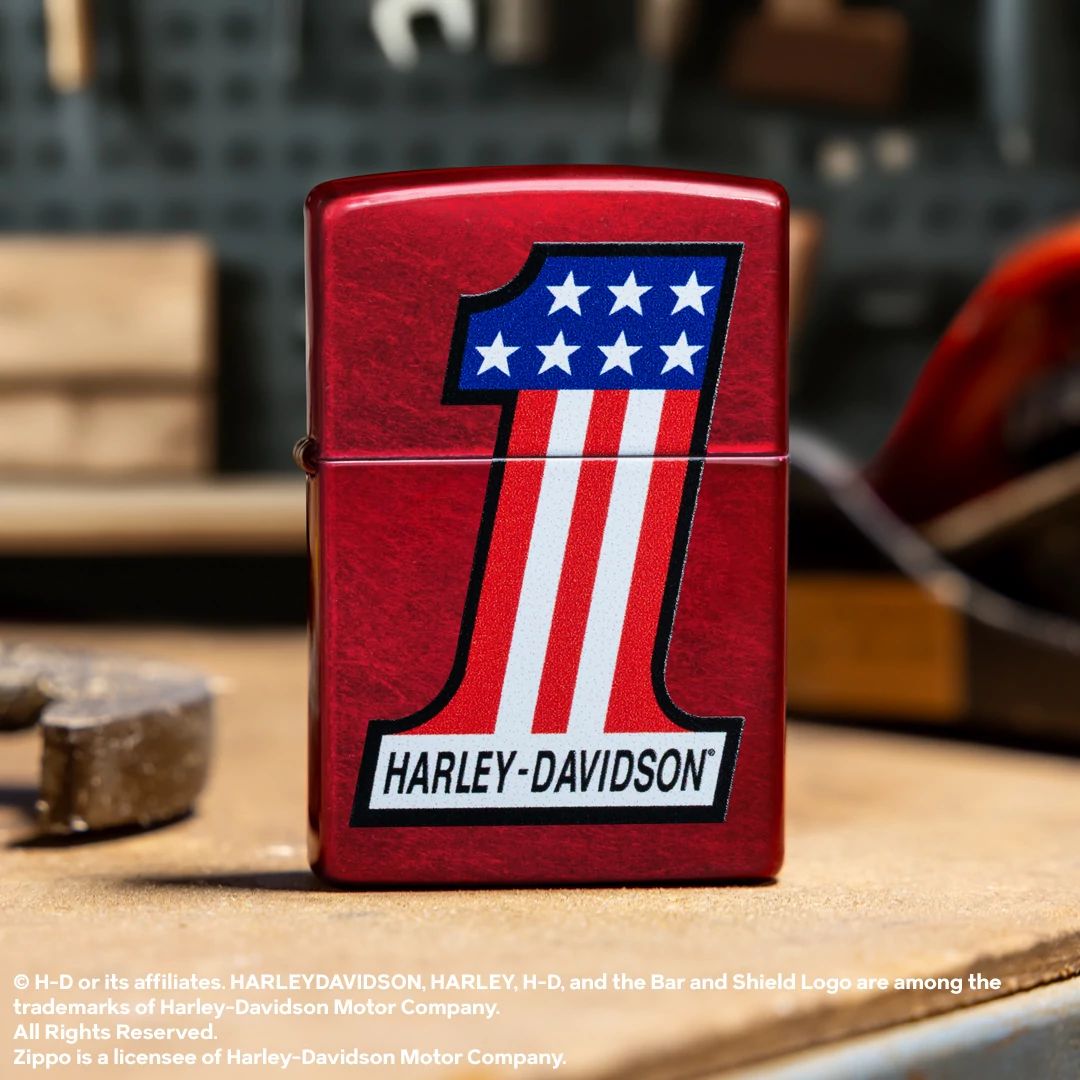 Lifestyle image of Zippo Harley-Davidson® #1 Logo Design Candy Apple Red Windproof Lighter standing next to tools on a workbench showing the front of the lighter. 

