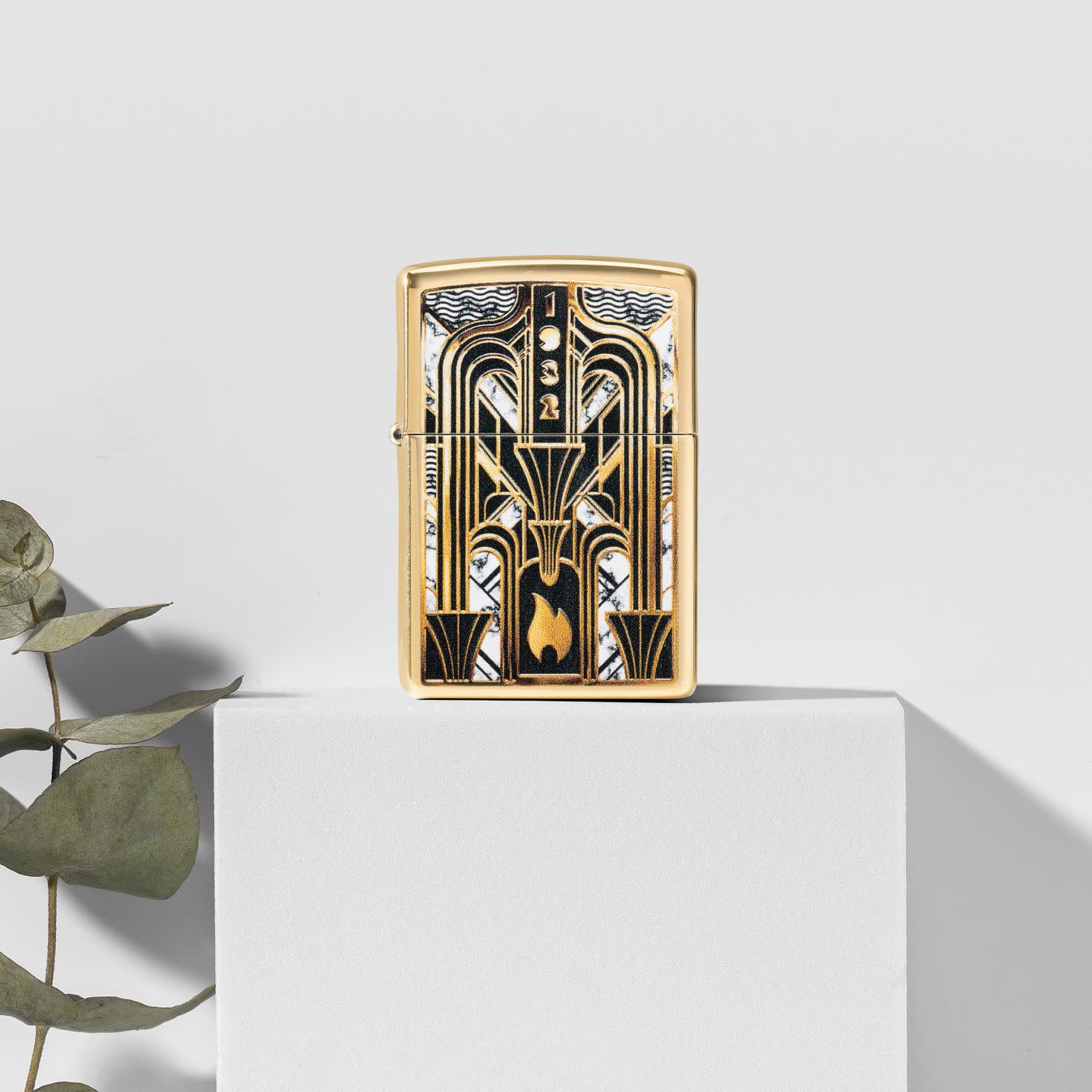 Lifestyle image of Zippo Art Deco Flame Design High Polish Brass Windproof Lighter standing on a white block with a green plant next to it on a white background.