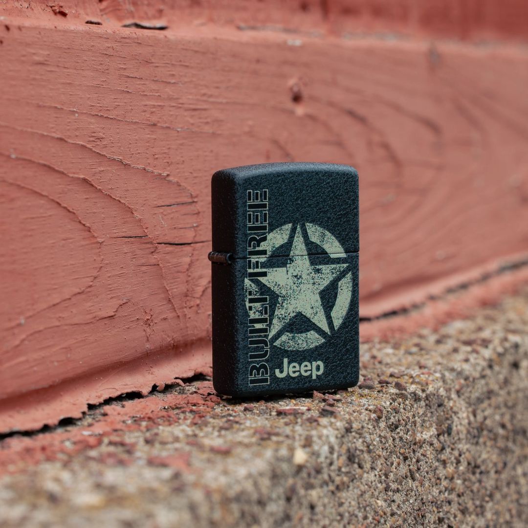 Lifestyle image of Zippo Jeep® Freedom Star Design Black Crackle Windproof Lighter standing at a 3/4 angle on a concrete ledge showing the front and hinge side of the lighter with a painted wooden background.

