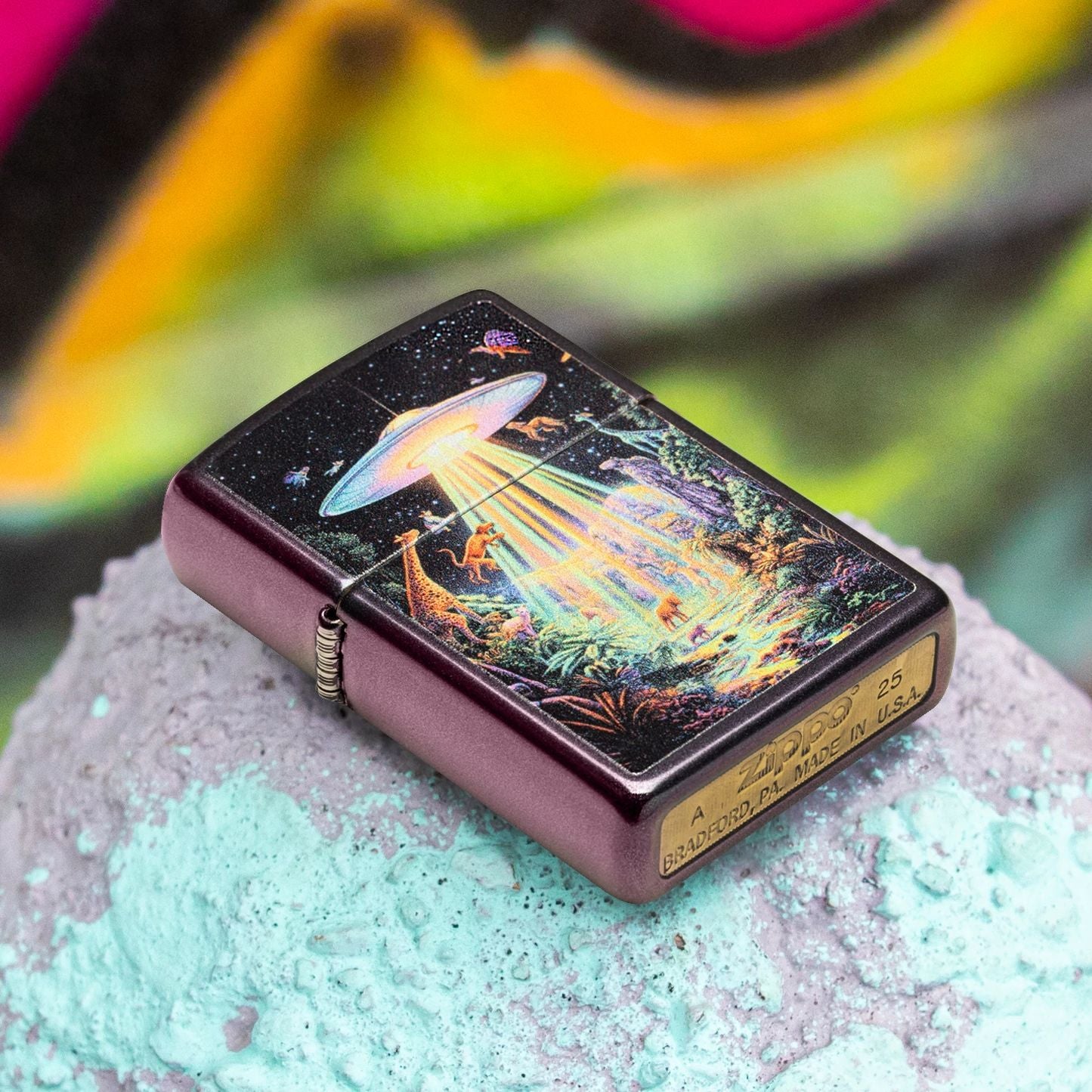 Lifestyle image of Zippo Animal Abduction Design Iridescent Windproof Lighter lying flat on the top of a concrete post while showing the front, hinge side, and bottom stamp of the lighter. The background is a bright graffiti covered wall.

