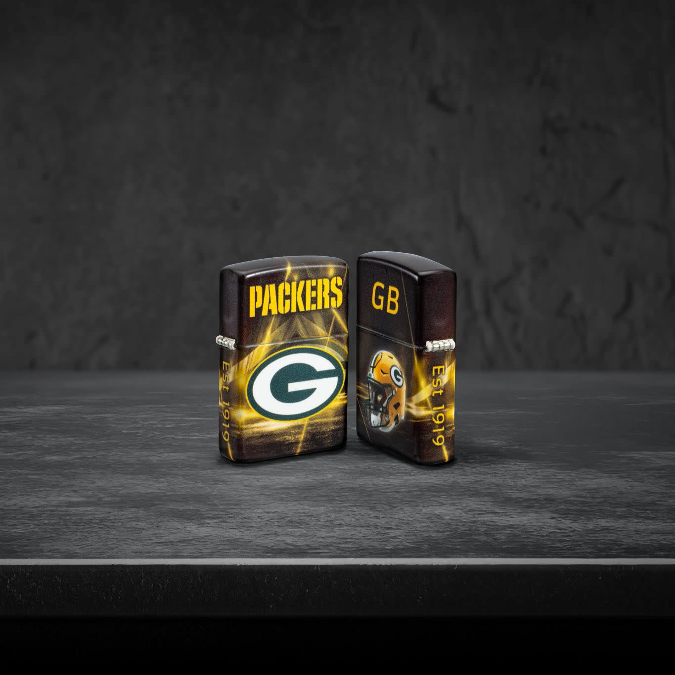 Lifestyle image of two Zippo NFL Green Bay Packers 540 Matte Windproof Lighters, one showing the front of the lighter and the other showing the back, standing on a dark grey surface.