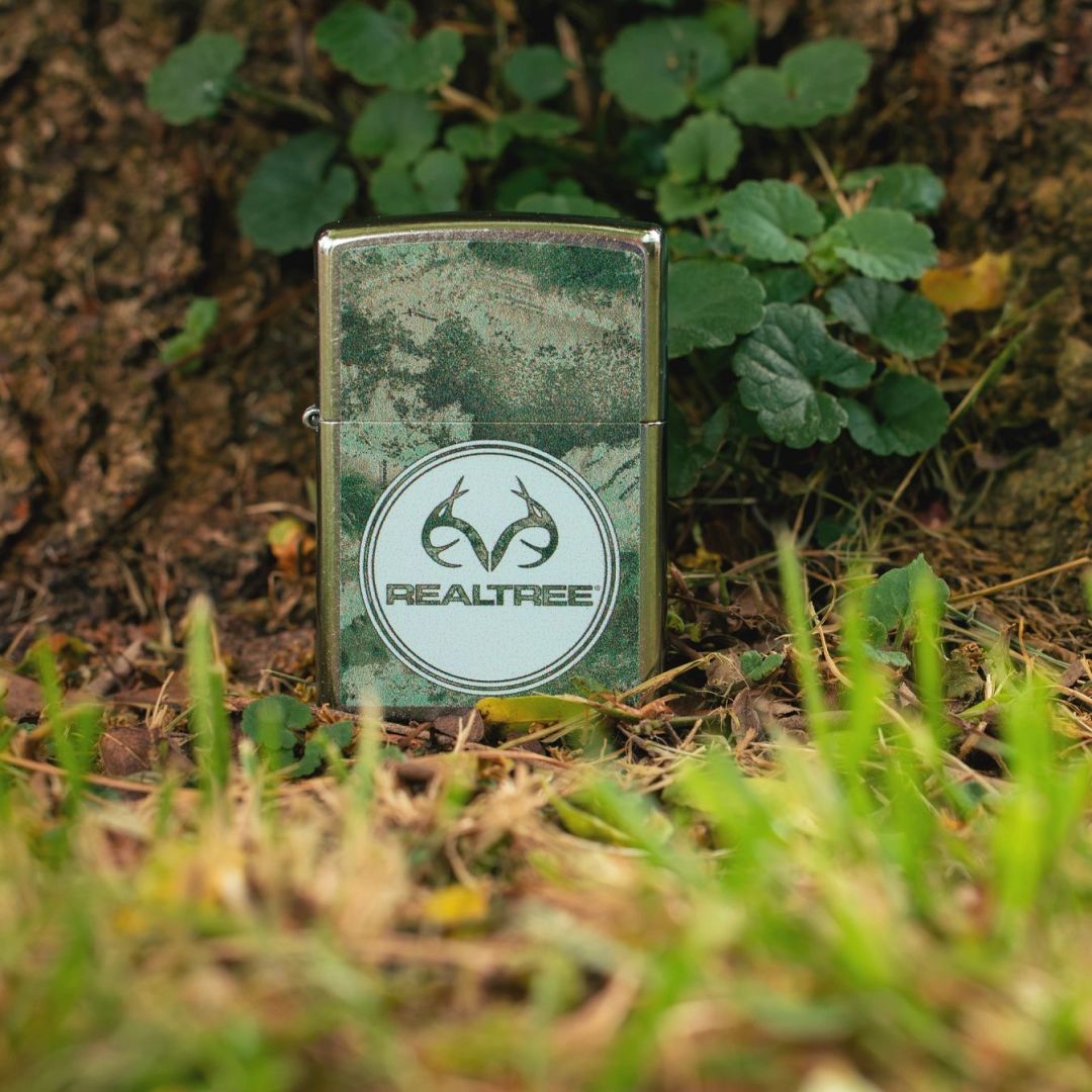 Lifestyle image of Zippo Realtree® XTREME Design Street Chrome Windproof Lighter standing on the ground showing the front of the lighter with the base of a tree as the background. 

