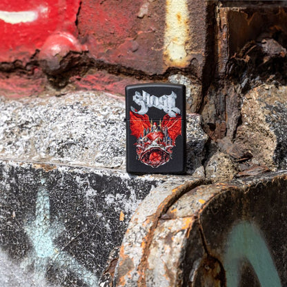 Lifestyle image of Zippo Ghost Prequelle Design Black Matte Windproof Lighter standing next to a graffiti-covered concrete structure showing the front of the lighter. 

