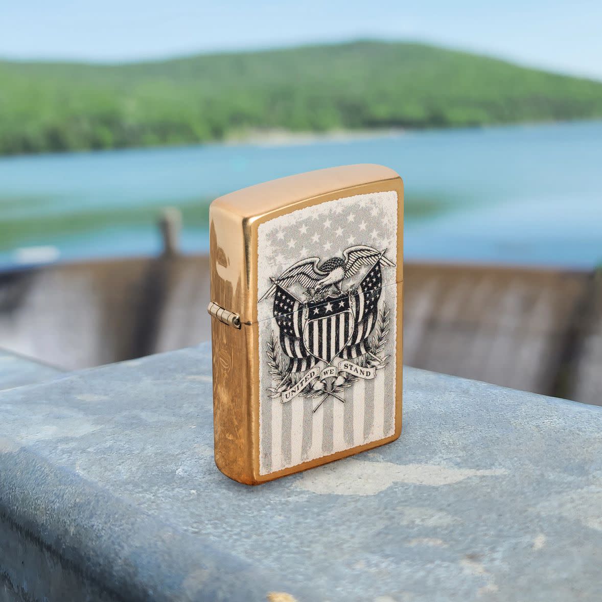 Lifestyle image of Zippo American Crest Design Brass Brushed Windproof Lighter standing on a metal container with water and hills in the background.