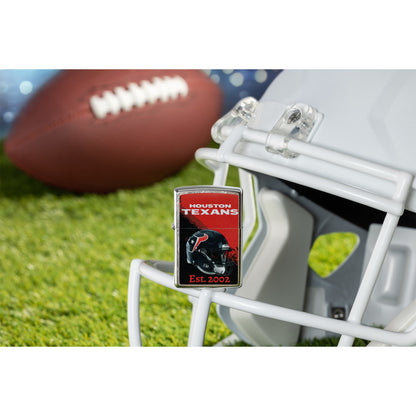 Lifestyle image of Zippo NFL Houston Texans Helmet Design Street Chrome Windproof Lighter standing on the facemask of a white helmet showing the front of the lighter with a football lying on a football field as the background.


