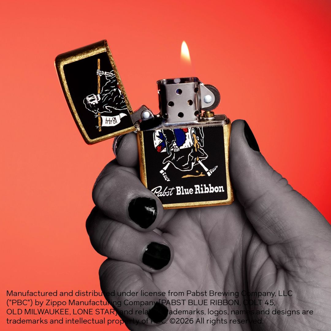 Lifestyle image of Zippo Pabst Blue Ribbon® Reaper Design Street Brass Windproof Lighter in hand open and lit showing the front of the lighter with a sunset-red ombre background. 

