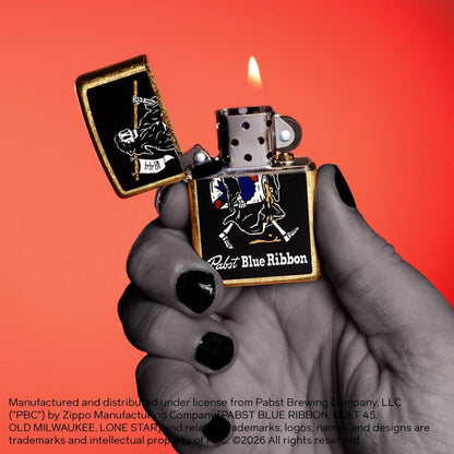 Lifestyle image of Zippo Pabst Blue Ribbon® Reaper Design Street Brass Windproof Lighter in hand open and lit showing the front of the lighter with a sunset-red ombre background. 

