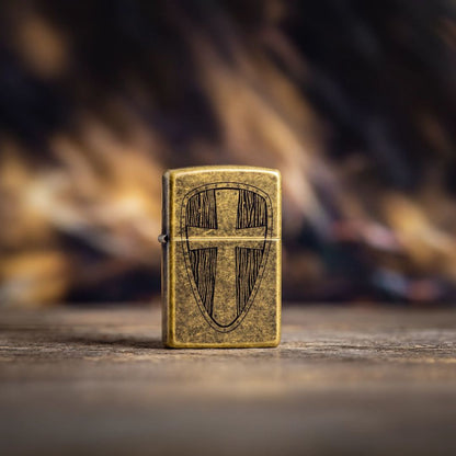 Lifestyle image of Zippo Medieval Shield Design Antique Brass Windproof Lighter standing on a dark stone floor showing the front of the lighter with a dark out of focus background.

