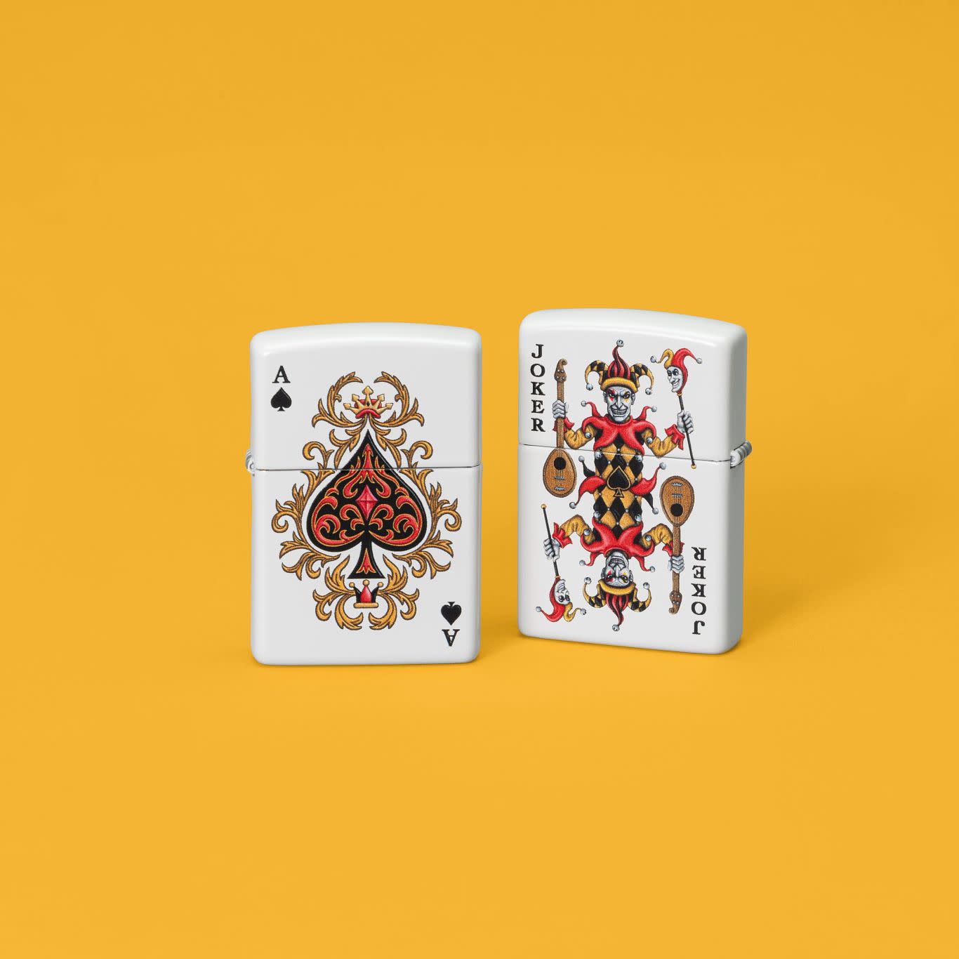 Lifestyle image of two Zippo Ace of Jokes 2-Sided White Matte Windproof Lighters set on a orange-yellow background.