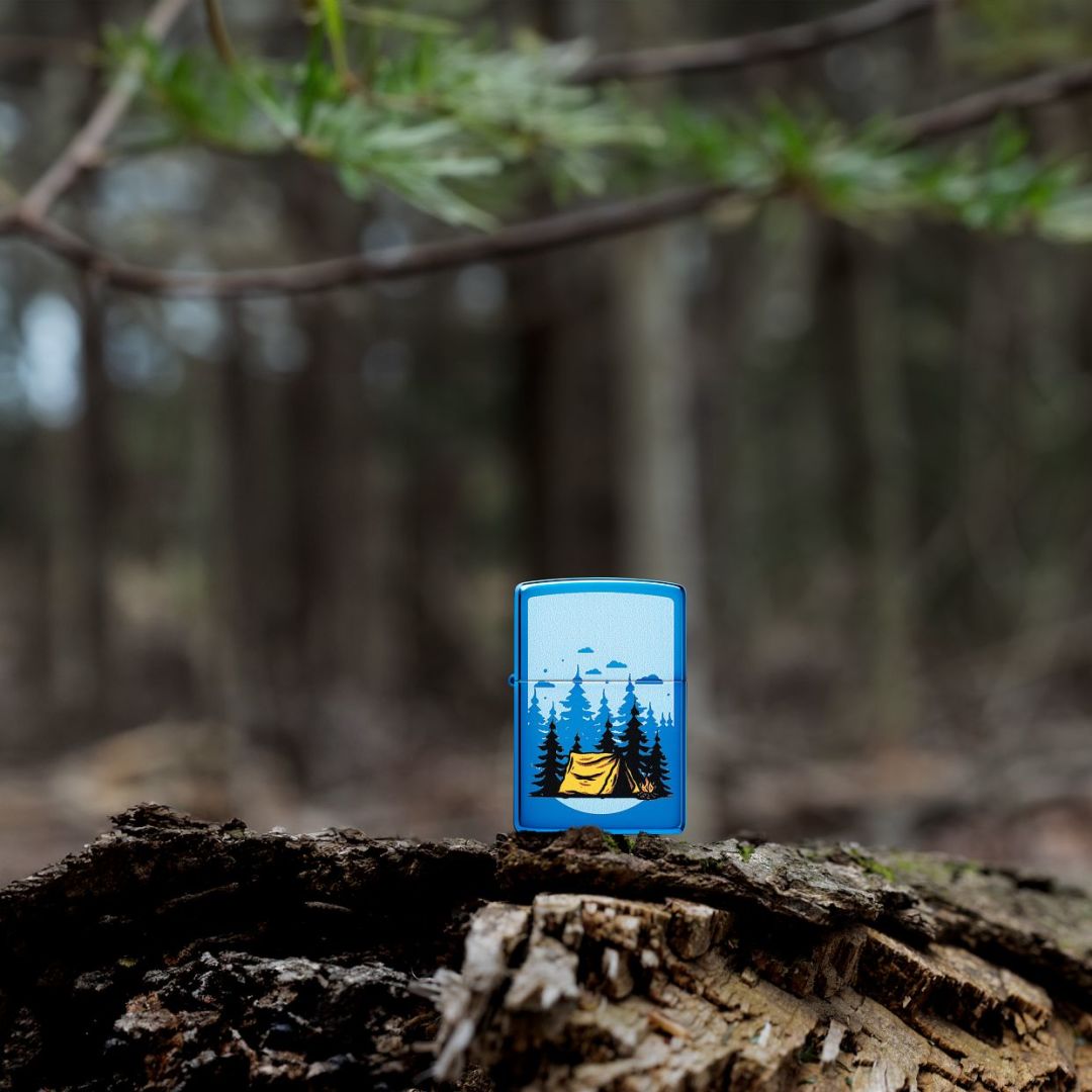 Lifestyle image of Zippo Forest Flicker Design High Polish Blue Windproof Lighter standing on a tree stump showing the front of the lighter with the forest as the background. 

