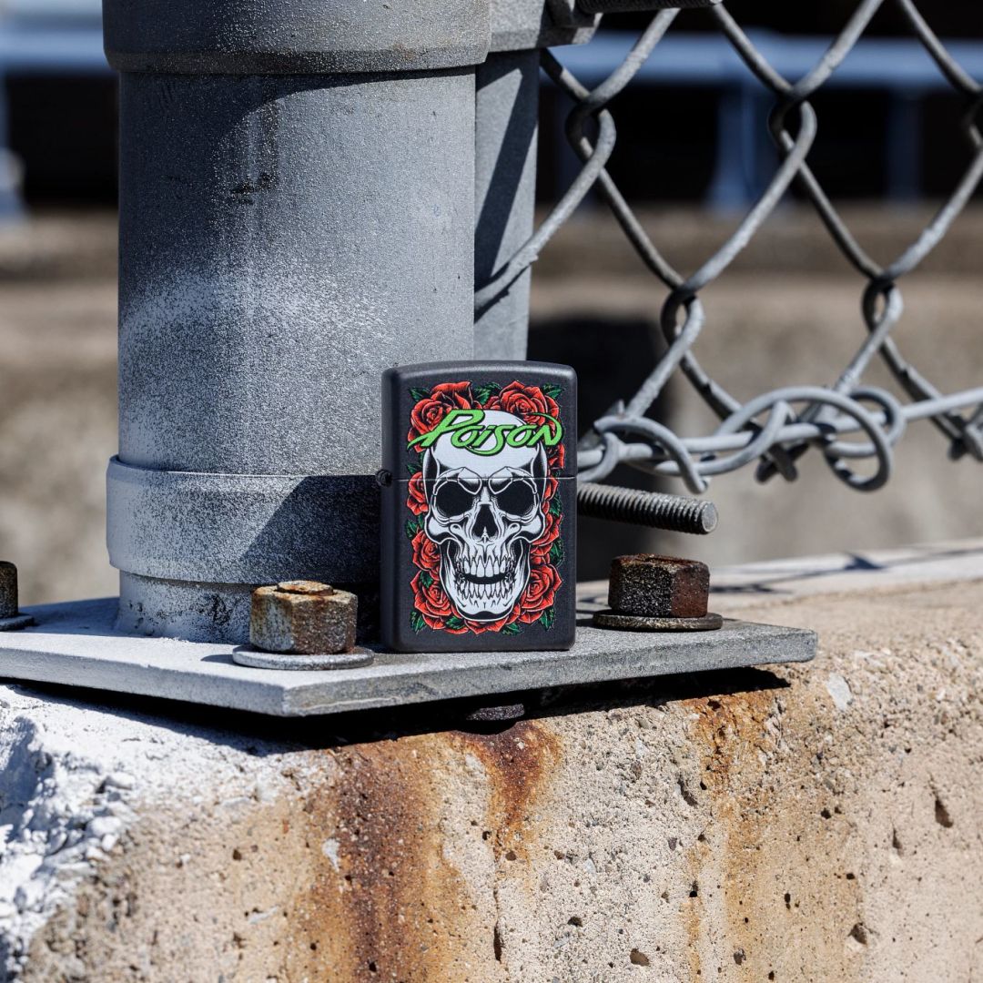 Lifestyle image of Zippo Poison Skull and Roses Design Black Matte Windproof Lighter standing at the base of a chain link fence post showing the front of the lighter with the post and fence as the background. 

