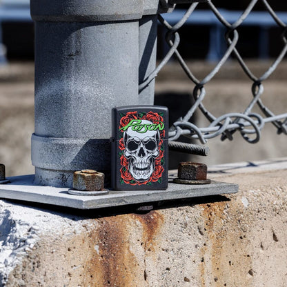 Lifestyle image of Zippo Poison Skull and Roses Design Black Matte Windproof Lighter standing at the base of a chain link fence post showing the front of the lighter with the post and fence as the background. 

