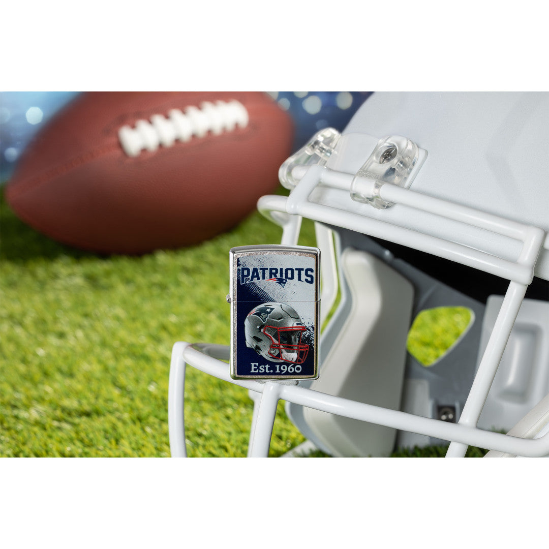Lifestyle image of Zippo NFL New England Patriots Helmet Design Street Chrome Windproof Lighter standing on the facemask of a white helmet showing the front of the lighter with a football lying on a football field as the background.

