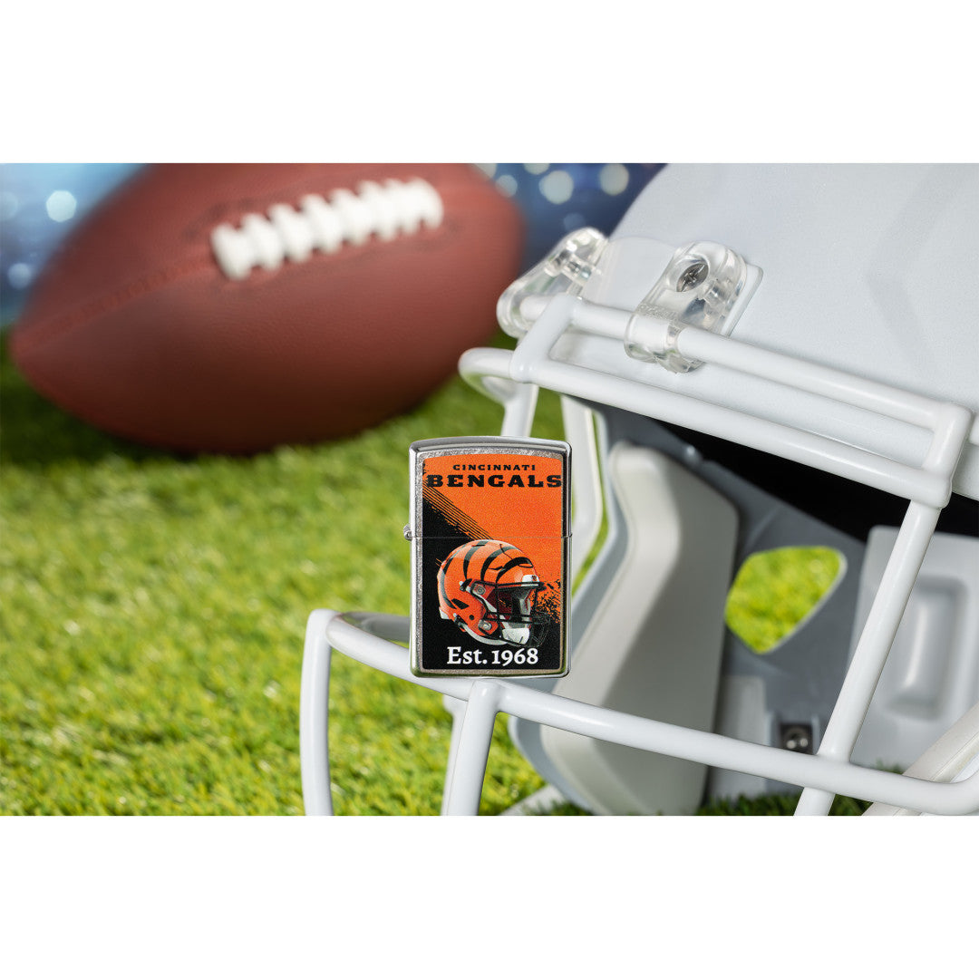 Lifestyle image of Zippo NFL Cincinnati Bengals Helmet Design Street Chrome Windproof Lighter standing on the facemask of a white helmet showing the front of the lighter with a football lying on a football field as the background.

