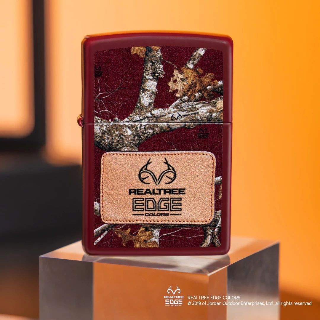 Lifestyle image of Zippo Realtree® Edge® Colors Design Merlot Windproof Lighter standing on a clear cube showing the front of the lighter with a light orange background.

