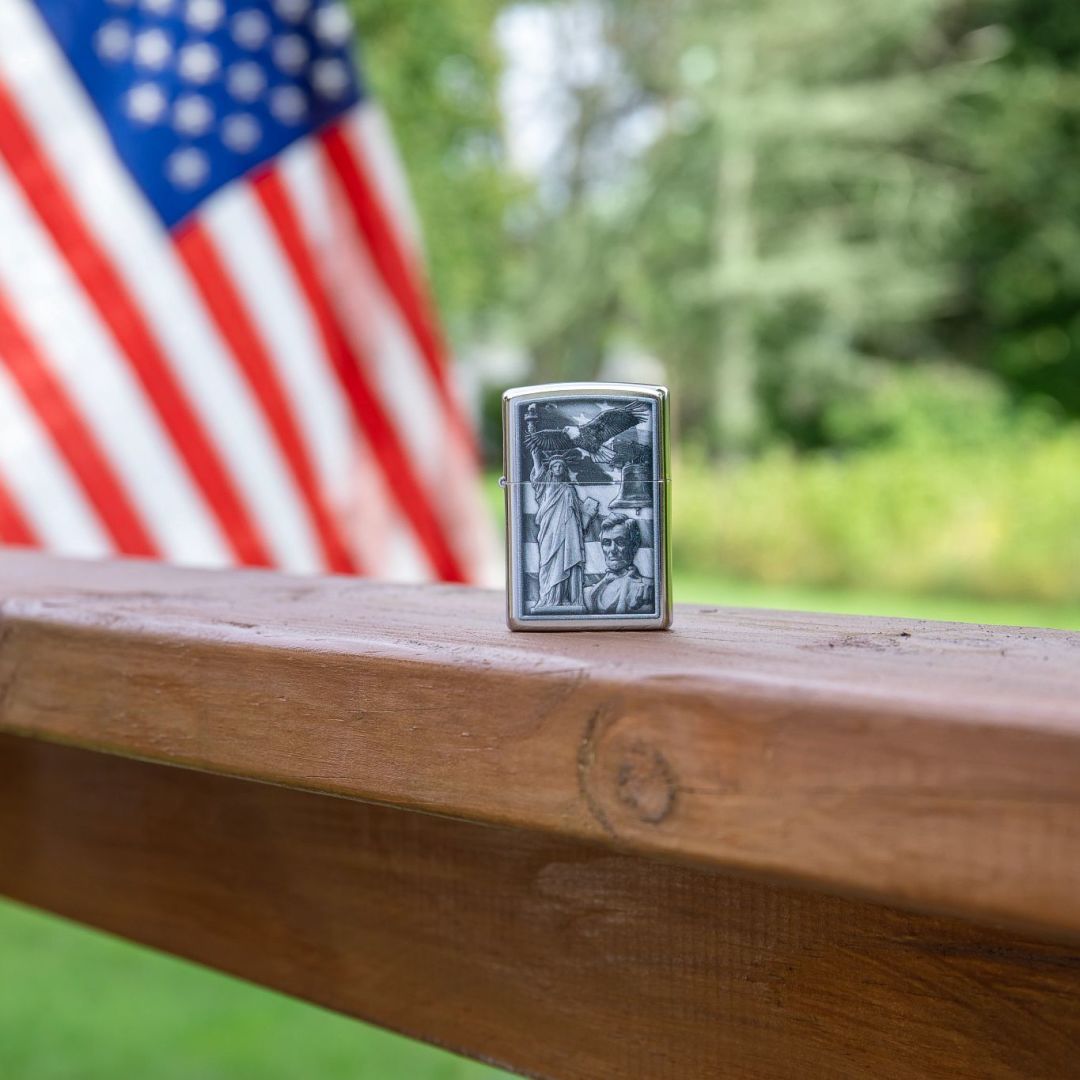 Lifestyle image of Zippo American Icons Design Street Chrome Windproof Lighter standing on a wooden railing showing the front of the lighter with a wooded backyard and an American flag as a background. 


