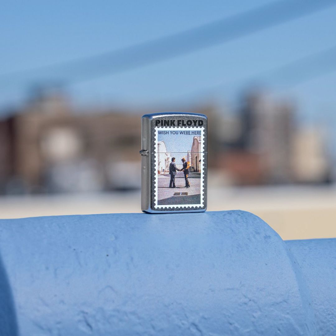 Lifestyle image of Zippo Pink Floyd Wish You Were Here Design Street Chrome Windproof Lighter standing on a light blue surface showing the front of the lighter with an out of focus city as the background. 

