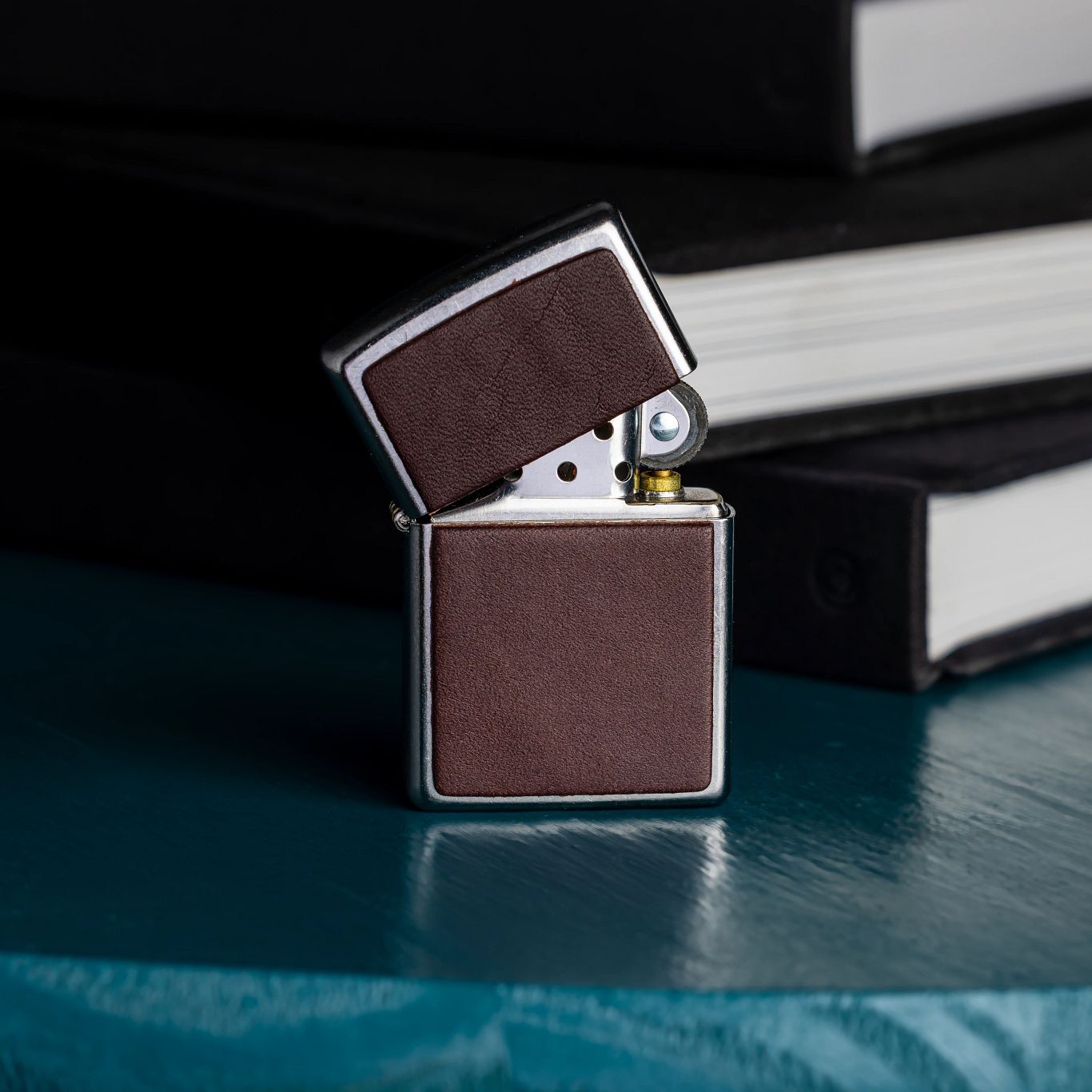 Lifestyle image of Zippo Brown Leather Emblem Street Chrome Windproof Lighter standing, slightly opened, on a desk next to three books. 

