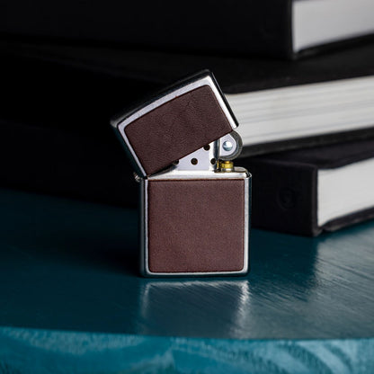 Lifestyle image of Zippo Brown Leather Emblem Street Chrome Windproof Lighter standing, slightly opened, on a desk next to three books. 

