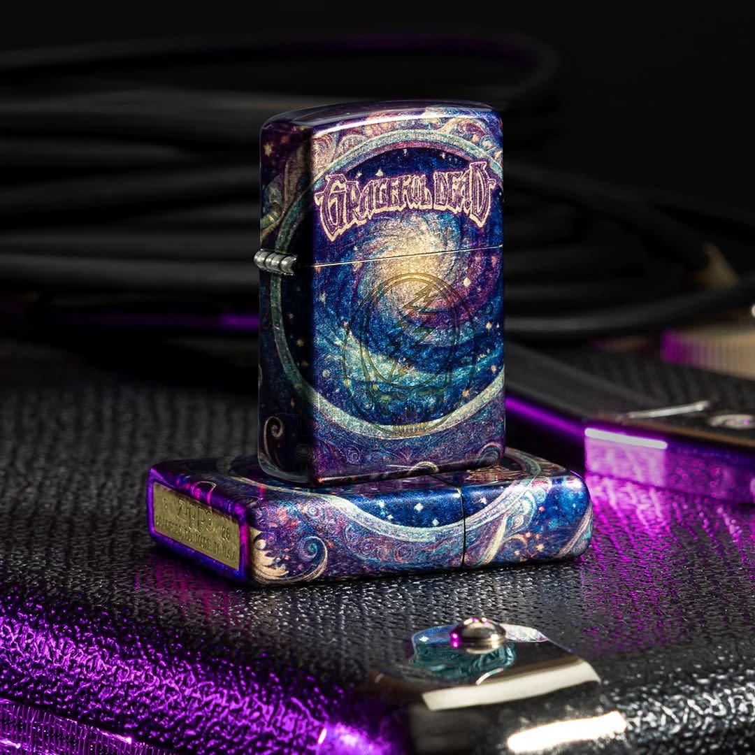 Lifestyle image of two Zippo Grateful Dead Limited Edition Stealie Design Tumbled Brass Windproof Lighters; one lying flat on an amp showing the non-hinge side and bottom stamp of the lighter while the other lighter is standing on top of the lighter that is lying flat showing the front and hinge side of the lighter with an amp and cords in the background while a purple light is shining on the amp. 

