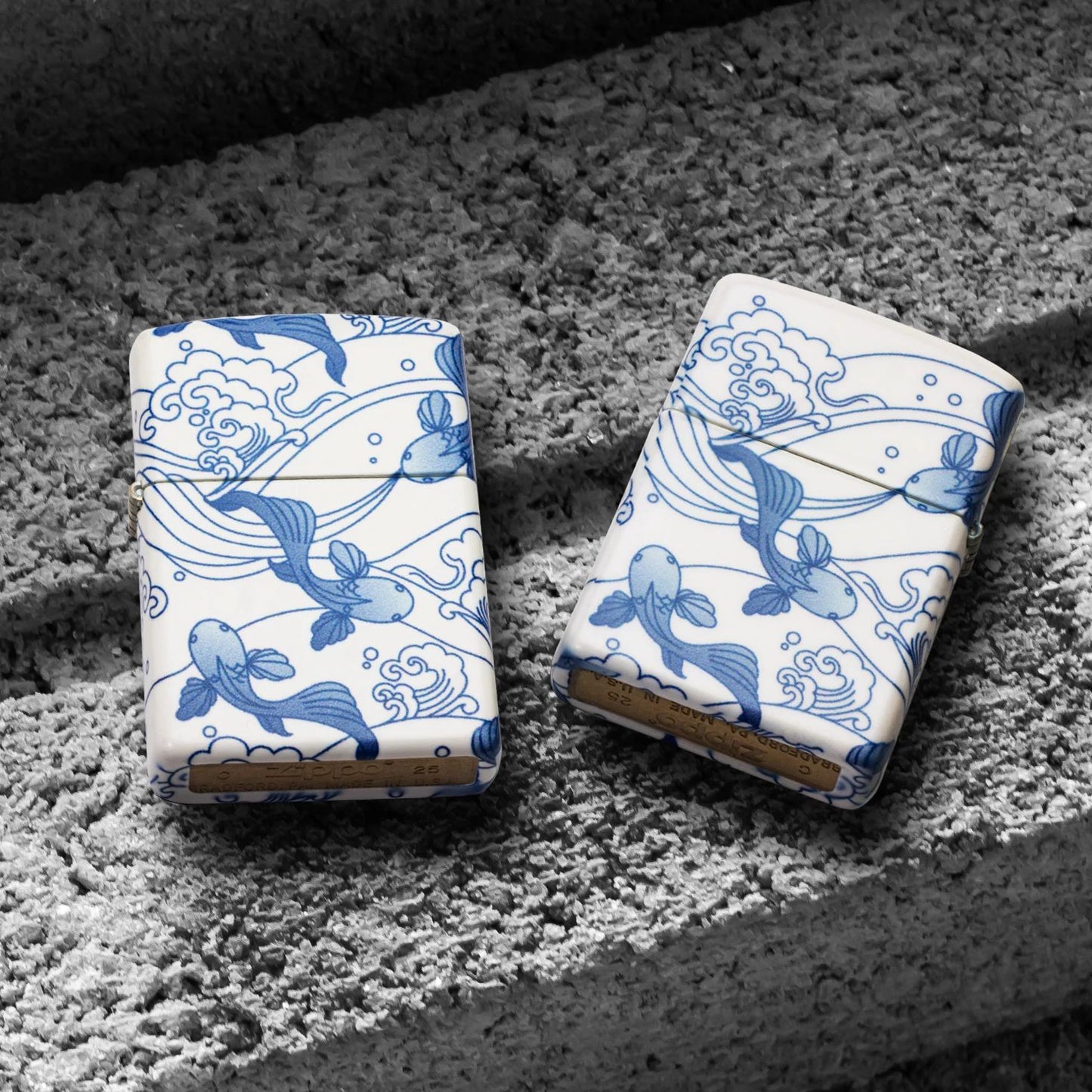 Lifestyle image of two Zippo Porcelain Koi Design 540 Matte Windproof Lighters; one showing the front of the lighter while the other is showing the back of the lighter, both lying on a cinder block. 

