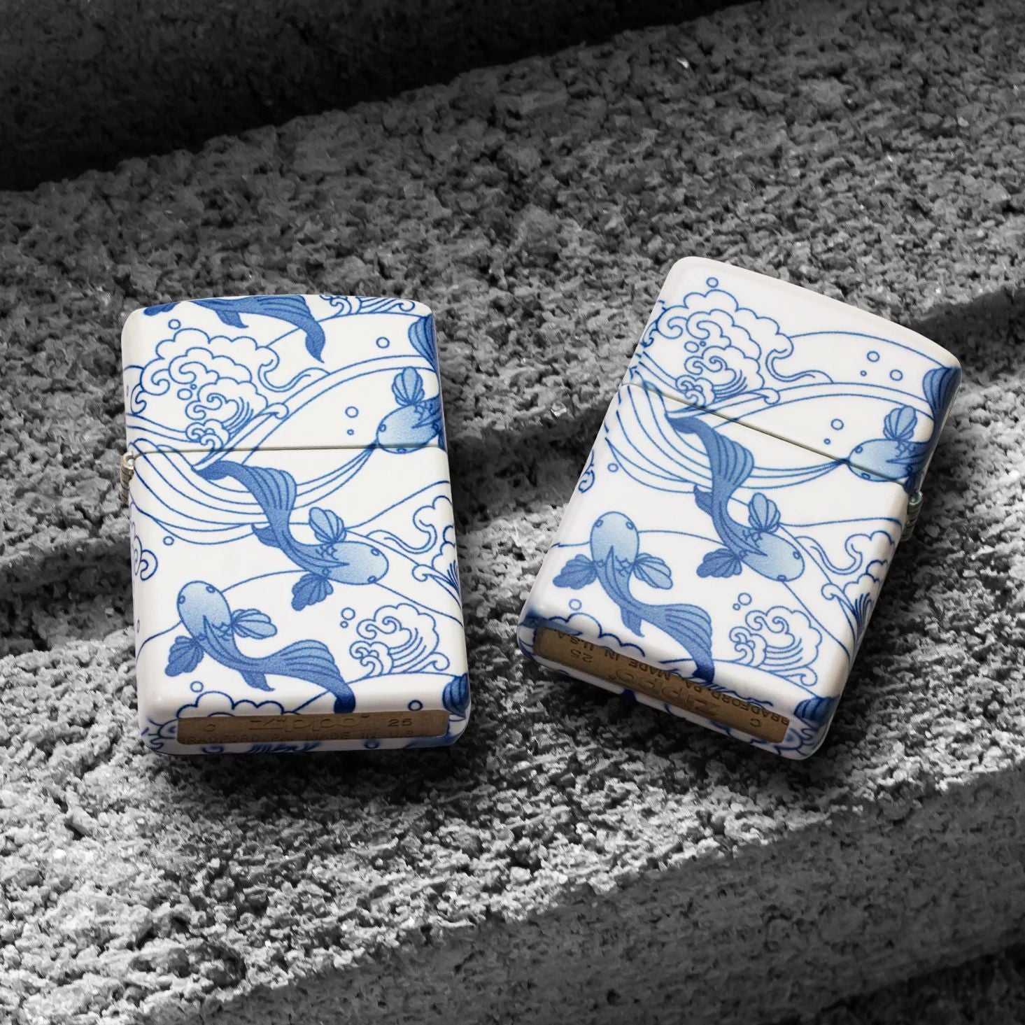 Lifestyle image of two Zippo Porcelain Koi Design 540 Matte Windproof Lighters; one showing the front of the lighter while the other is showing the back of the lighter, both lying on a cinder block. 

