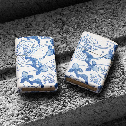 Lifestyle image of two Zippo Porcelain Koi Design 540 Matte Windproof Lighters; one showing the front of the lighter while the other is showing the back of the lighter, both lying on a cinder block. 

