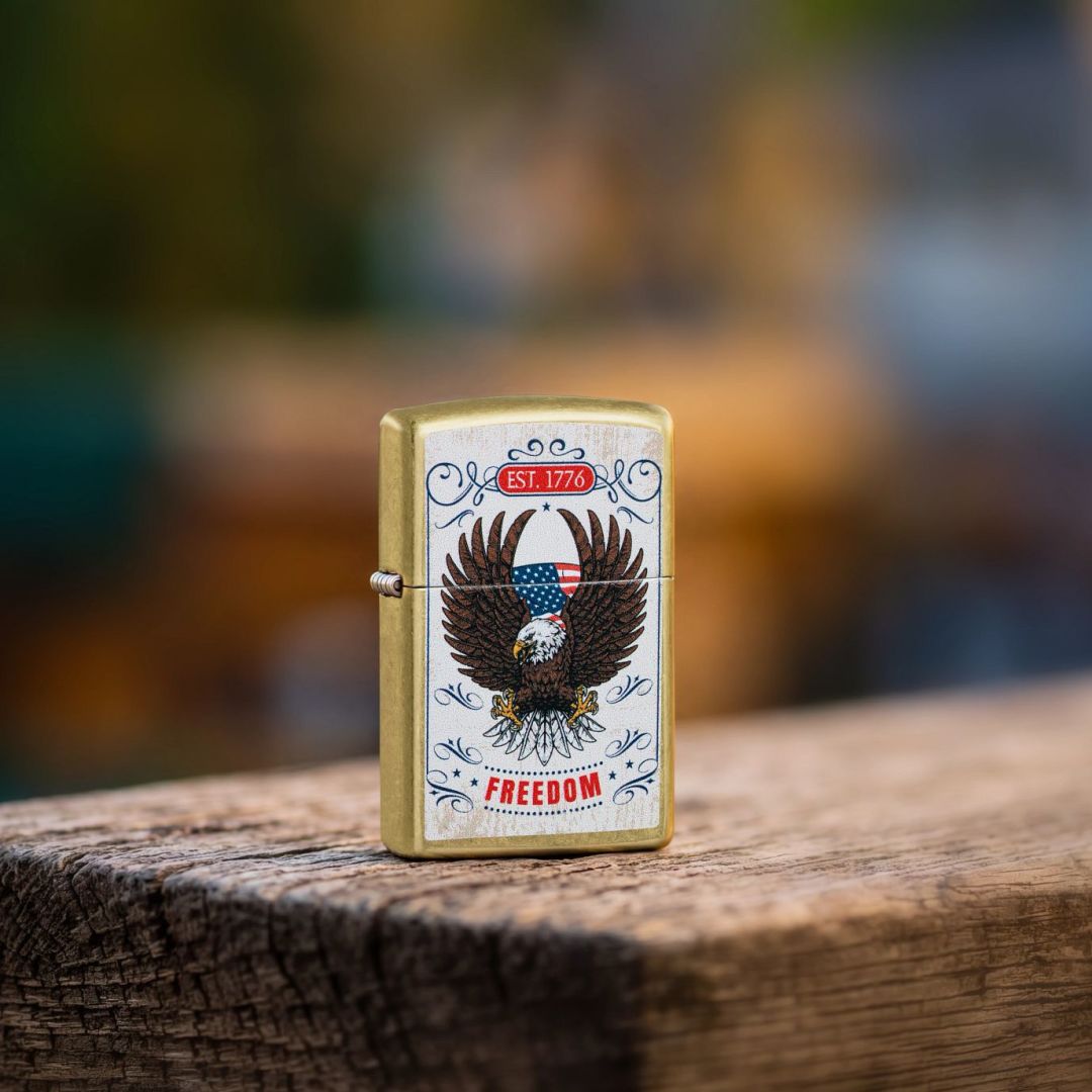 Lifestyle image of Zippo Soaring Freedom Design Street Brass Windproof Lighter standing on a wooden surface showing the front and hinge side of the lighter with an out of focus outdoor background. 

