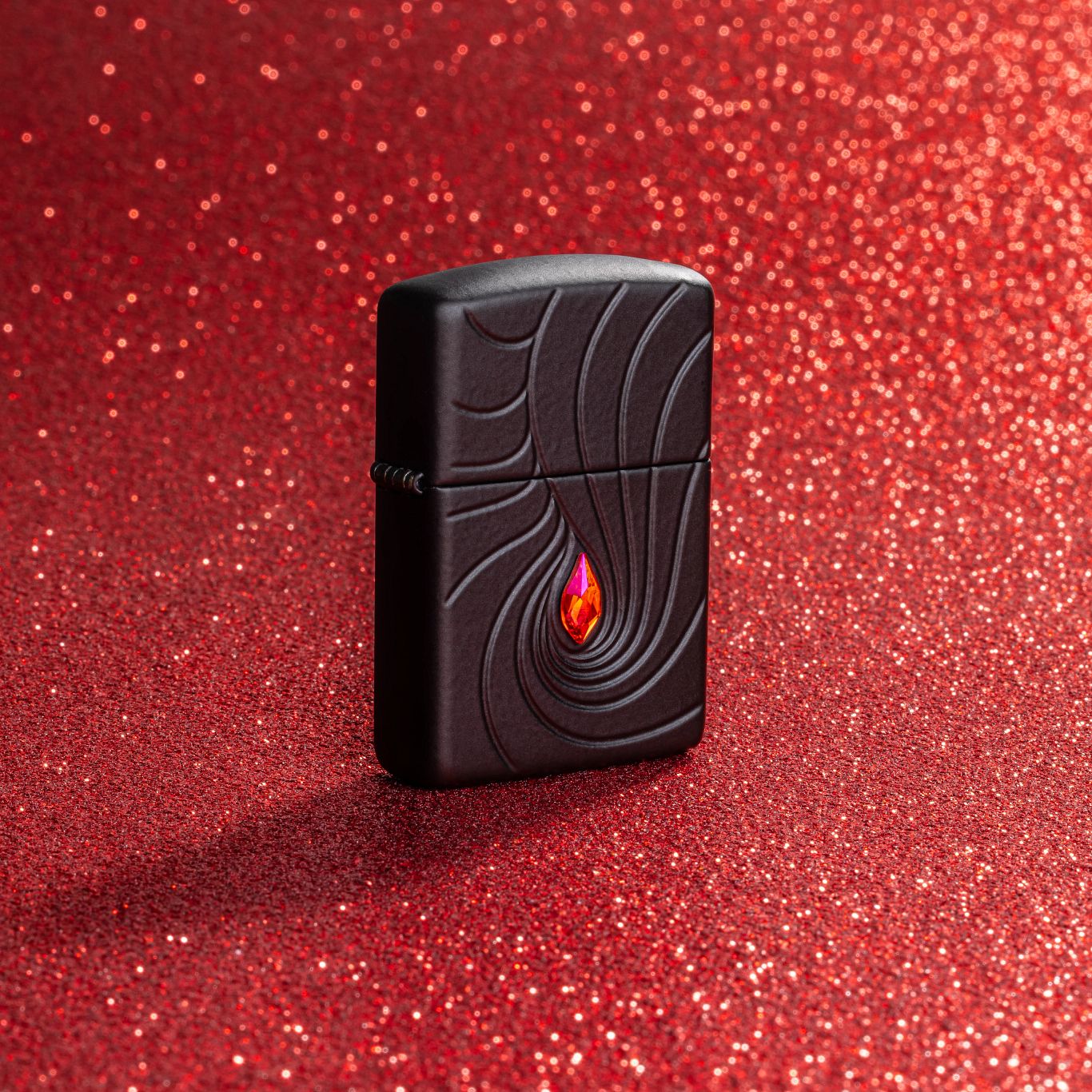 Lifestyle image of Zippo Crystal Waves Design Armor® Black Matte Windproof Lighter standing with a red glitter background.


