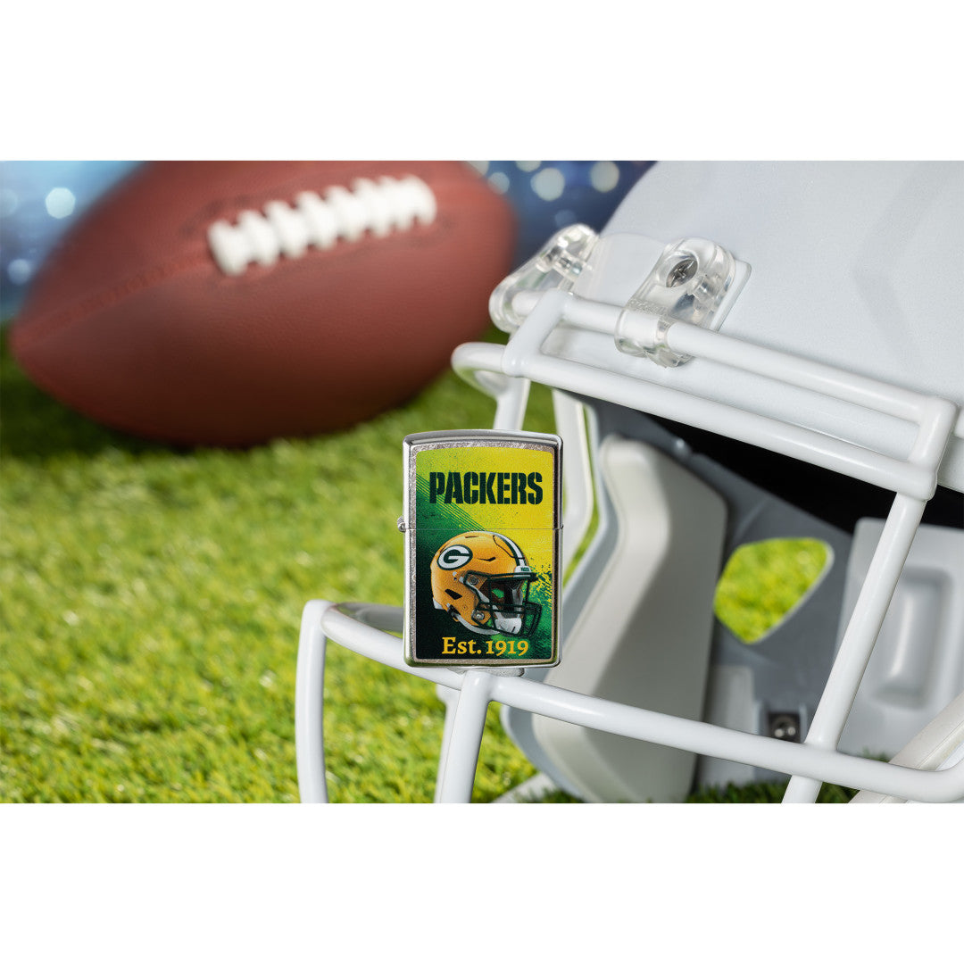 Lifestyle image of Zippo NFL Green Bay Packers Helmet Design Street Chrome Windproof Lighter standing on the facemask of a white helmet showing the front of the lighter with a football lying on a football field as the background.

