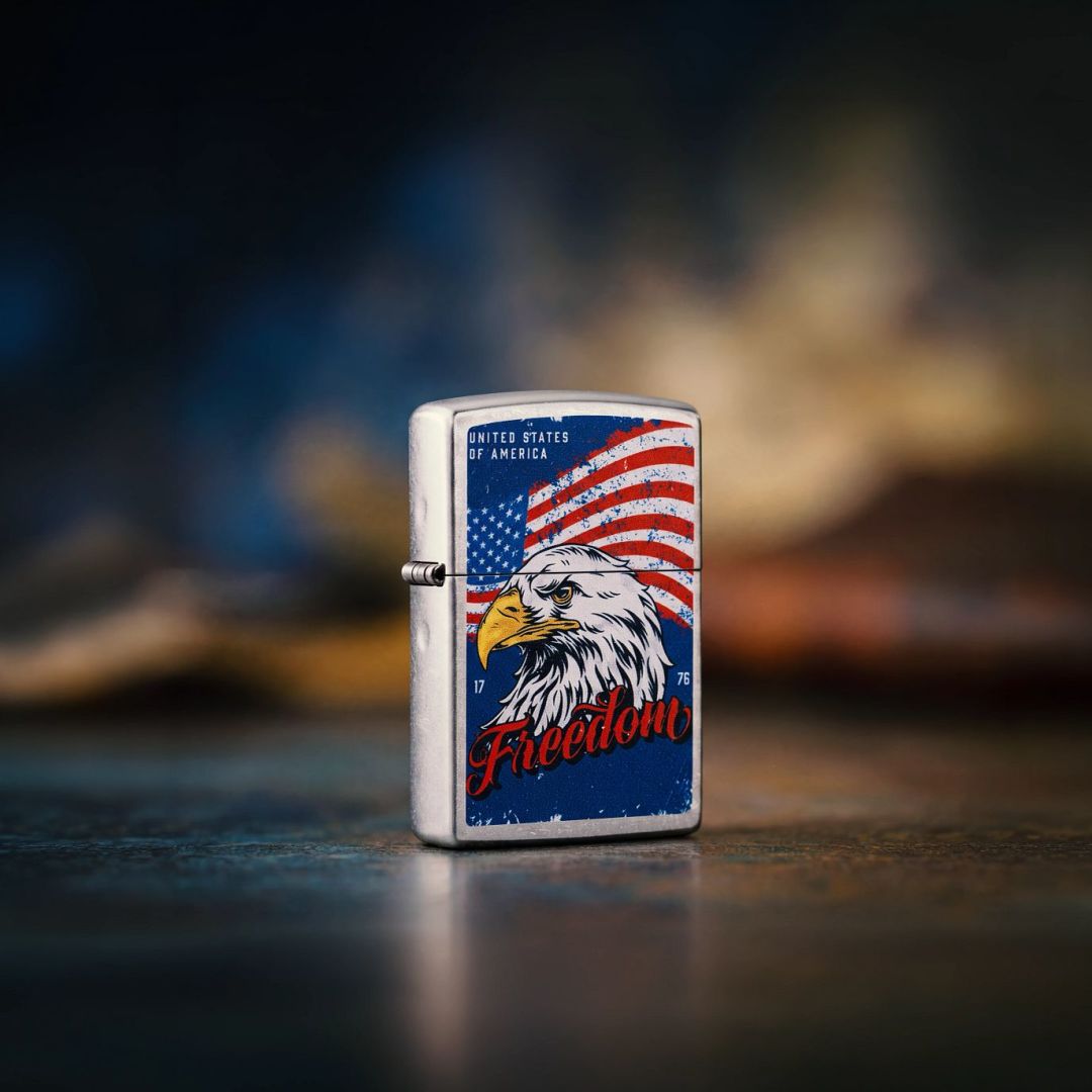 Lifestyle image of Zippo Freedom Eagle Design Street Chrome Windproof Lighter standing on a stone surface showing the front and hinge side of the lighter with a dark muted red, white and blue background. 

