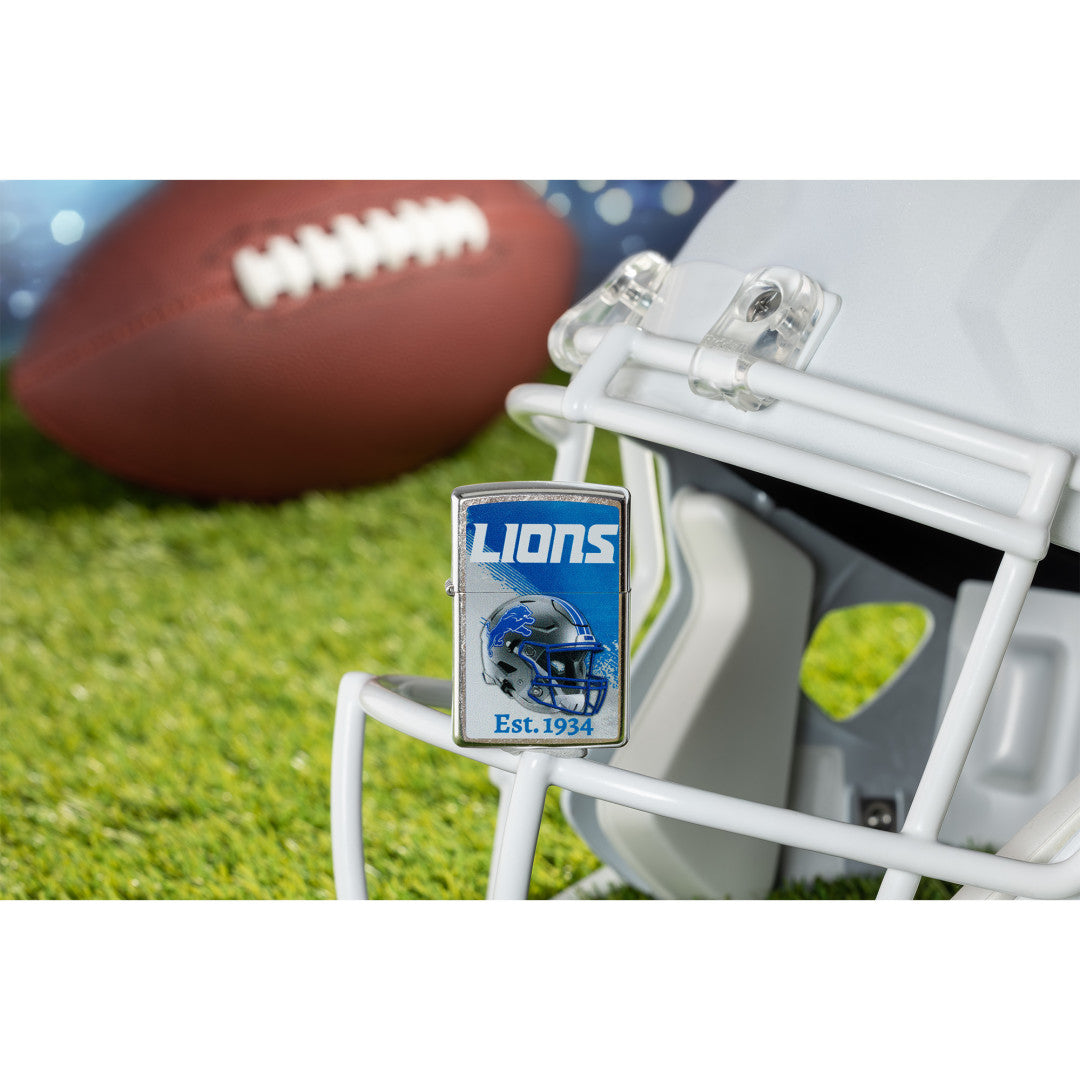 Lifestyle image of Zippo NFL Detroit Lions Helmet Design Street Chrome Windproof Lighter standing on the facemask of a white helmet showing the front of the lighter with a football lying on a football field as the background.

