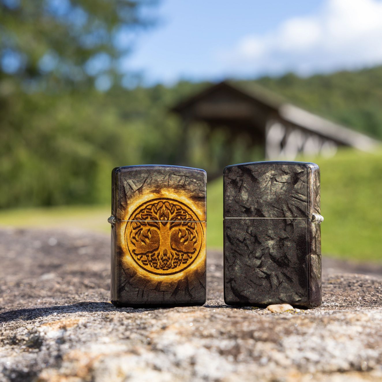 Lifestyle image of two Zippo Gleaming Tree of Life Design 540 Tumbled Brass Windproof Lighters; one showing the front of the lighter and the other showing the back and hinge side of the lighter, both standing on a boulder with a wooden covered bridge in a forest setting as the background.