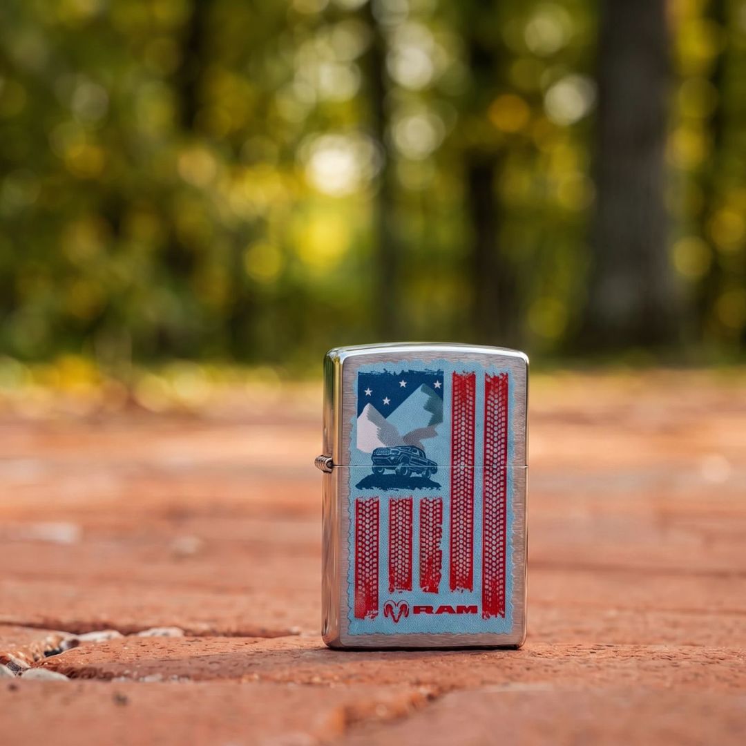 Lifestyle image of Zippo RAM Flag Design Brushed Chrome Windproof Lighter standing on a brick road showing the front of the lighter with an out of focus nature background. 

