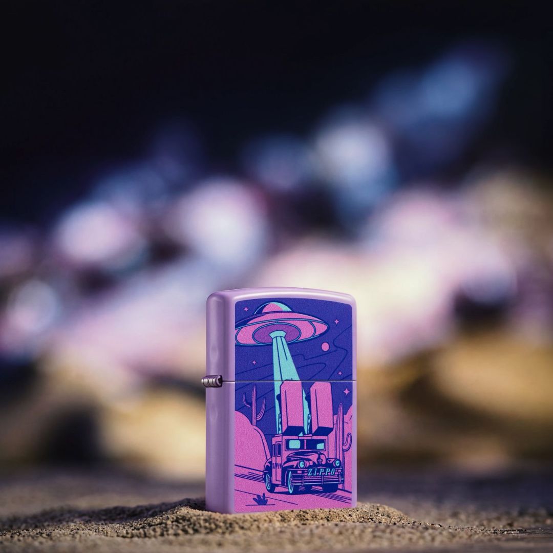 Lifestyle image of Zippo Car Chase Design Smoky Lavender Windproof Lighter standing in the sand showing the front and hinge side of the lighter with an out of focus lit-up background.

