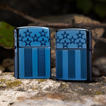 Lifestyle image of two Zippo Stars and Stripes Design Armor® High Polish Blue Windproof Lighters; one showing the front of the lighter and the other showing the back of the lighter, both standing on a rock with green leaves and twigs in the background. 

